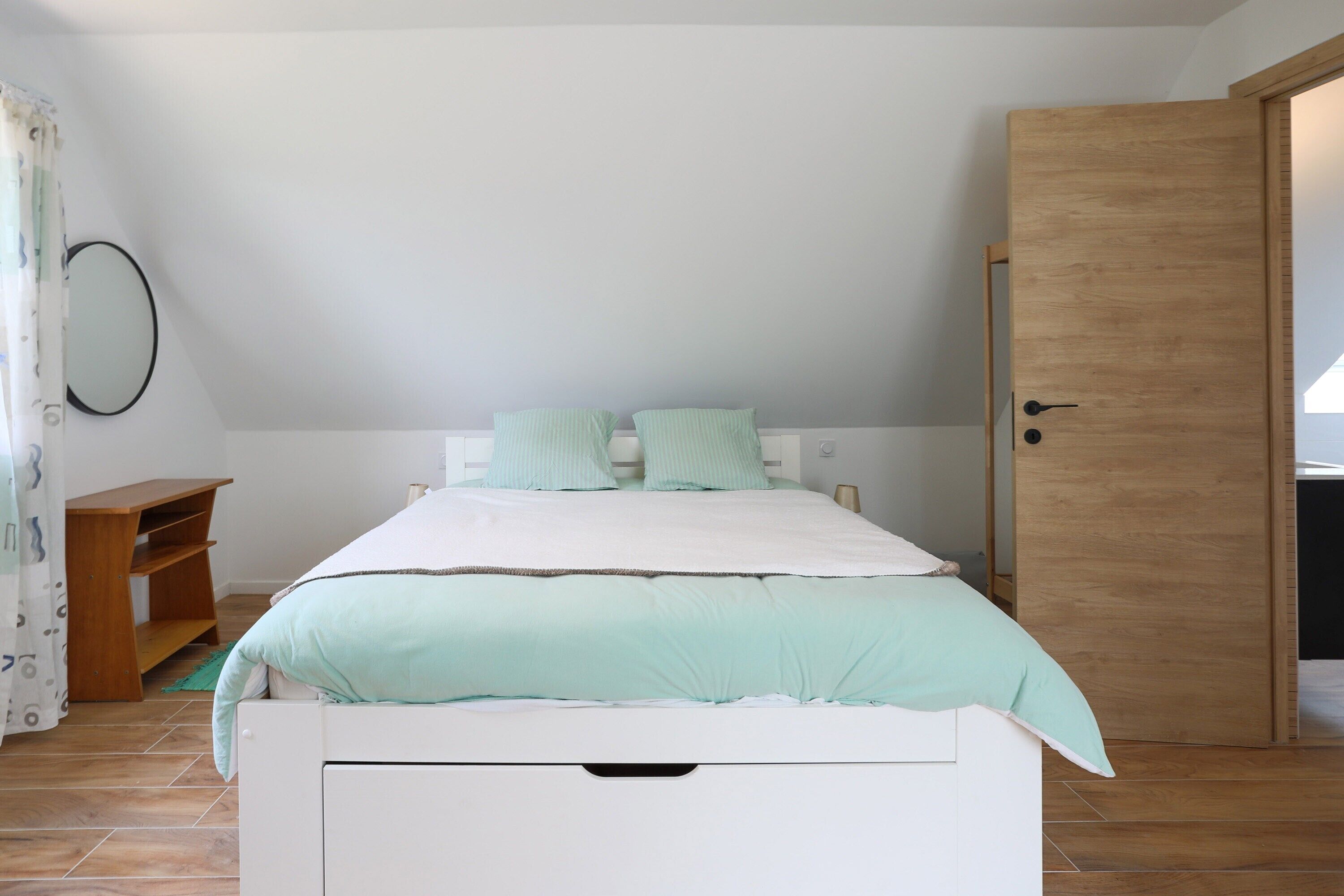 A bedroom with a bed, a wooden door, a small wooden shelf, and a wall-mounted mirror.