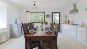 Un coin repas avec une table en bois dressée pour quatre personnes, entourée de chaises en osier, d’un luminaire suspendu et offrant une vue sur la verdure à l’extérieur.