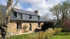 Une maison en pierre avec un toit en pente, une cheminée et un petit porche à l’avant, doté de sièges pour l’extérieur.