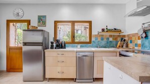 A modern kitchen with a refrigerator, dishwasher, wooden cabinets, a window with a view, and a clock on the wall.
