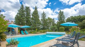 A swimming pool with lounge chairs and umbrellas, surrounded by trees and a stone building.