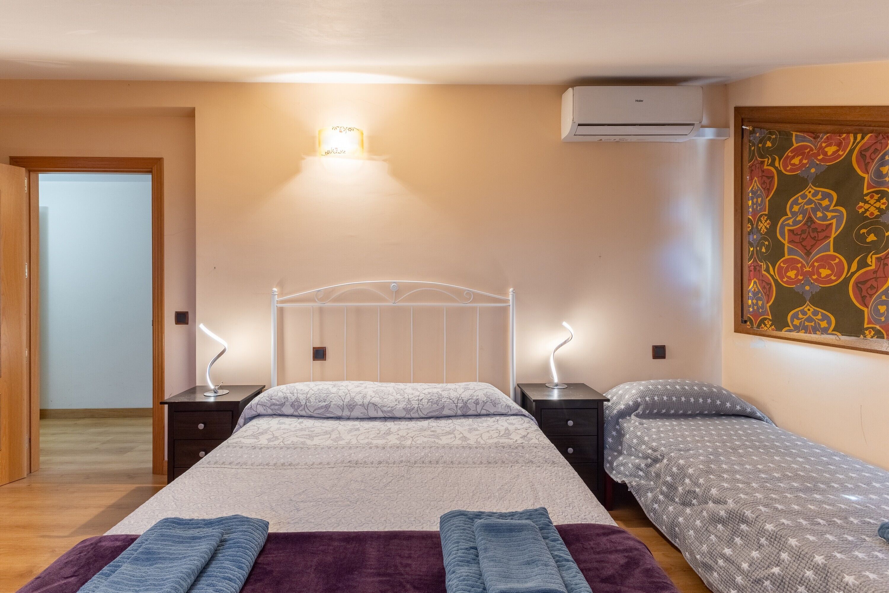 Un dormitorio con dos camas, una mesita de noche con lámpara, aire acondicionado e iluminación en la pared.