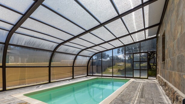 Una piscina coperta con un involucro in vetro, pavimento in legno e vista sull'esterno.