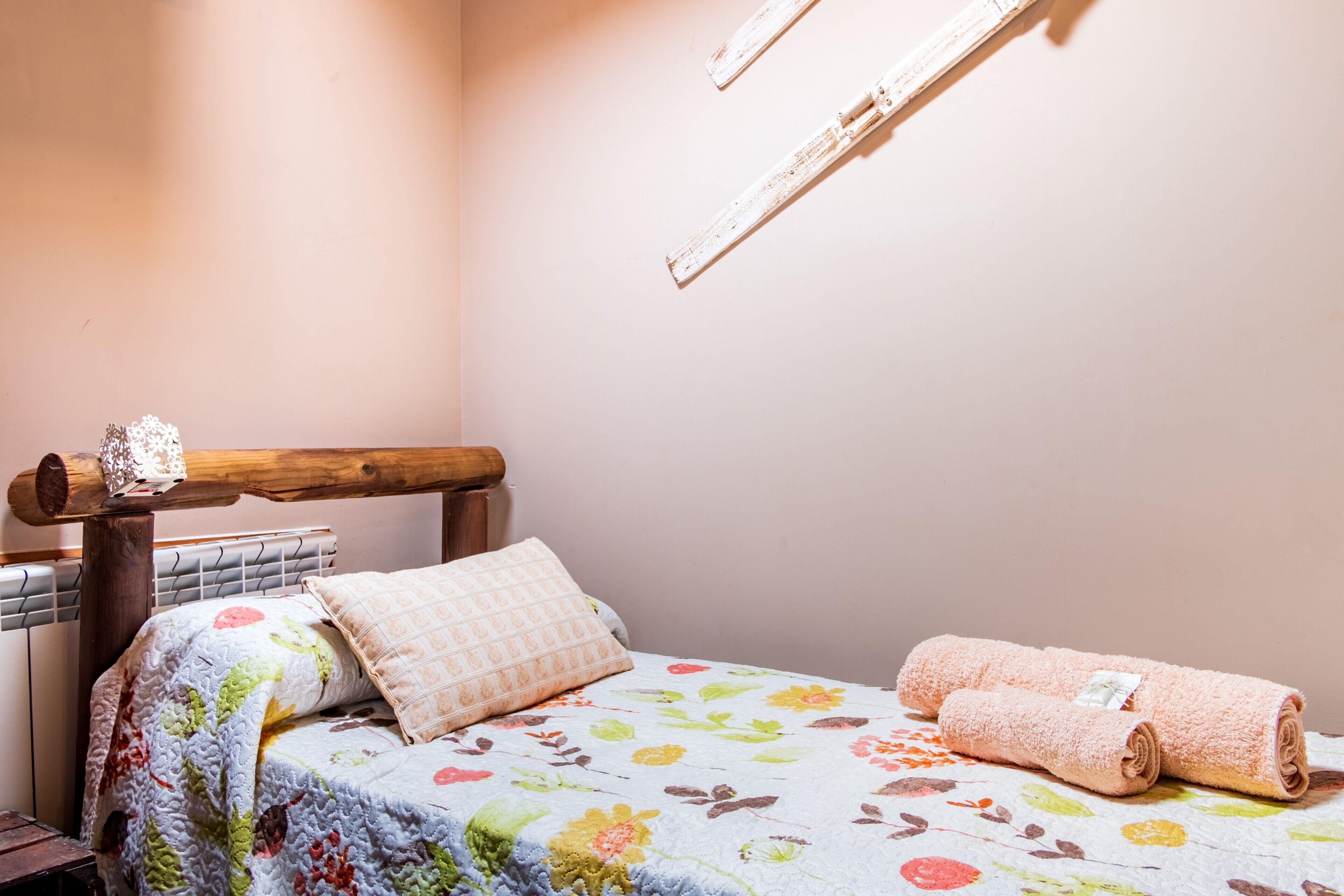 A bed with a floral quilt, a wooden headboard, a rolled towel, and a wall-mounted light fixture.
