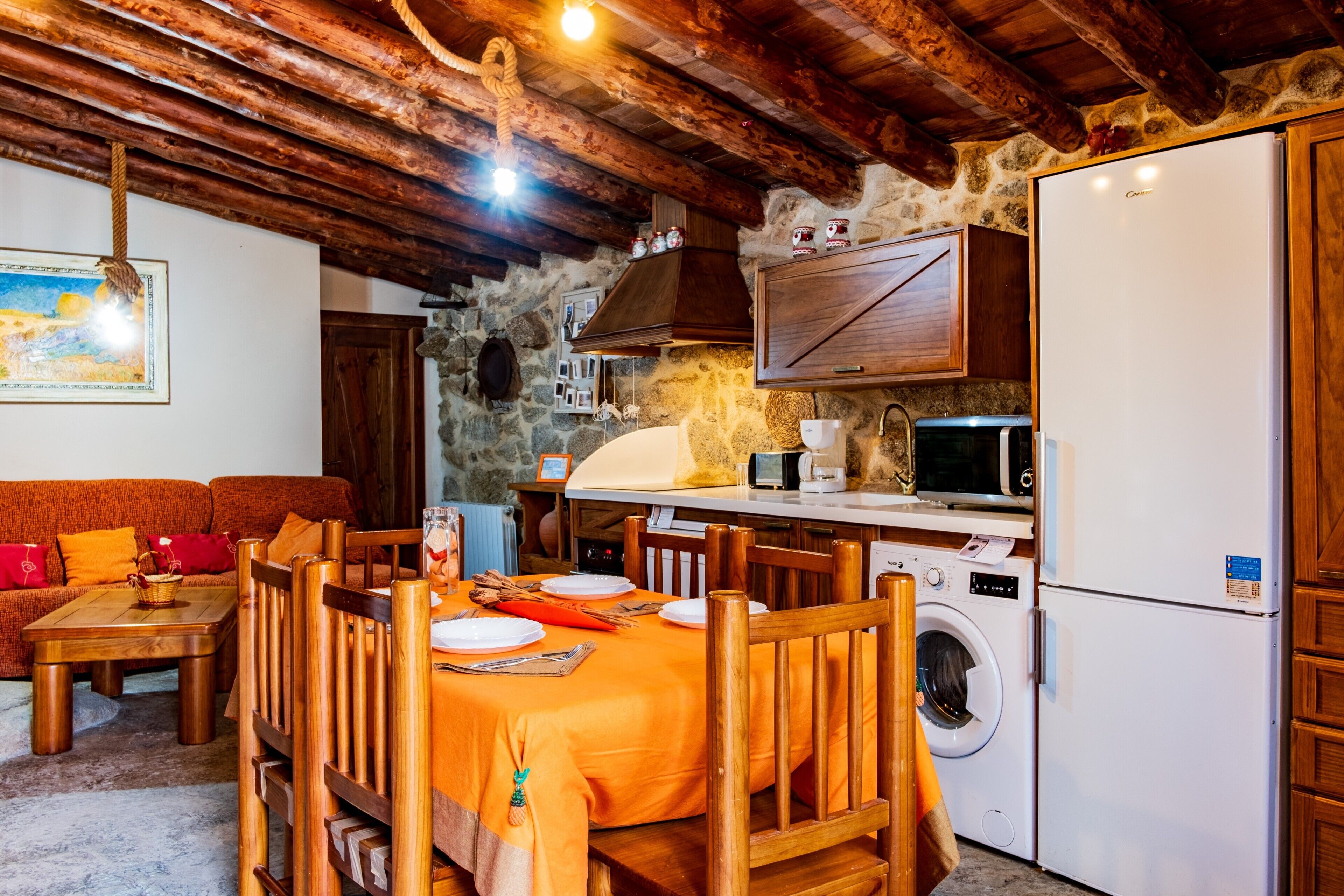 Une cuisine rustique avec un mur en pierre, des poutres en bois au plafond, une table à manger recouverte d’une nappe orange, des chaises en bois, un lave-linge et un réfrigérateur blanc.