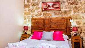 Un lit avec des oreillers roses et gris, une tête de lit en bois et un mur en pierre.