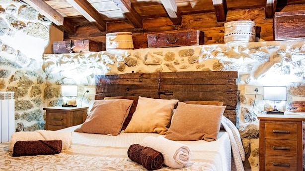 Ein Schlafzimmer mit Steinwänden, einem hölzernen Kopfteil, einem Bett mit beigefarbener und brauner Bettwäsche sowie einem Nachttisch aus Holz mit einer Lampe.