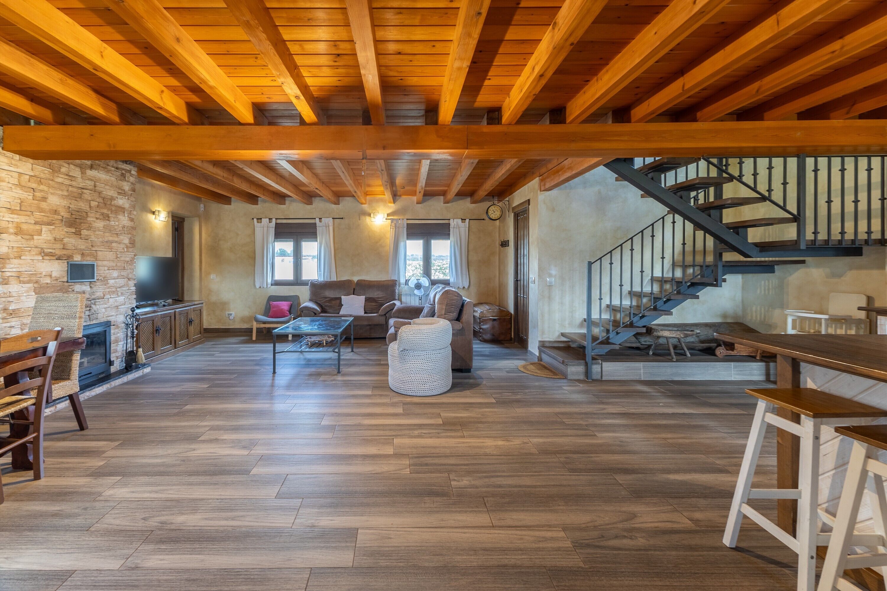 A spacious living area with a wooden ceiling, stone wall, and a staircase leading to an upper level.