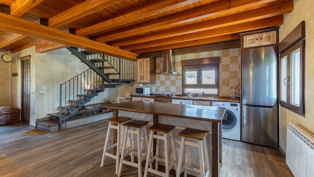A spacious kitchen with wooden beams, a central island with stools, a stainless steel refrigerator, and a washing machine. - Paraje Navazos – Rural House in Sierra de Gredos with Pool and Mountain Views (Gavilanes)