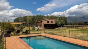 A swimming pool with a tiled deck, a covered seating area, and a stone building with a pitched roof. - Paraje Navazos – Rural House in Sierra de Gredos with Pool and Mountain Views (Gavilanes)
