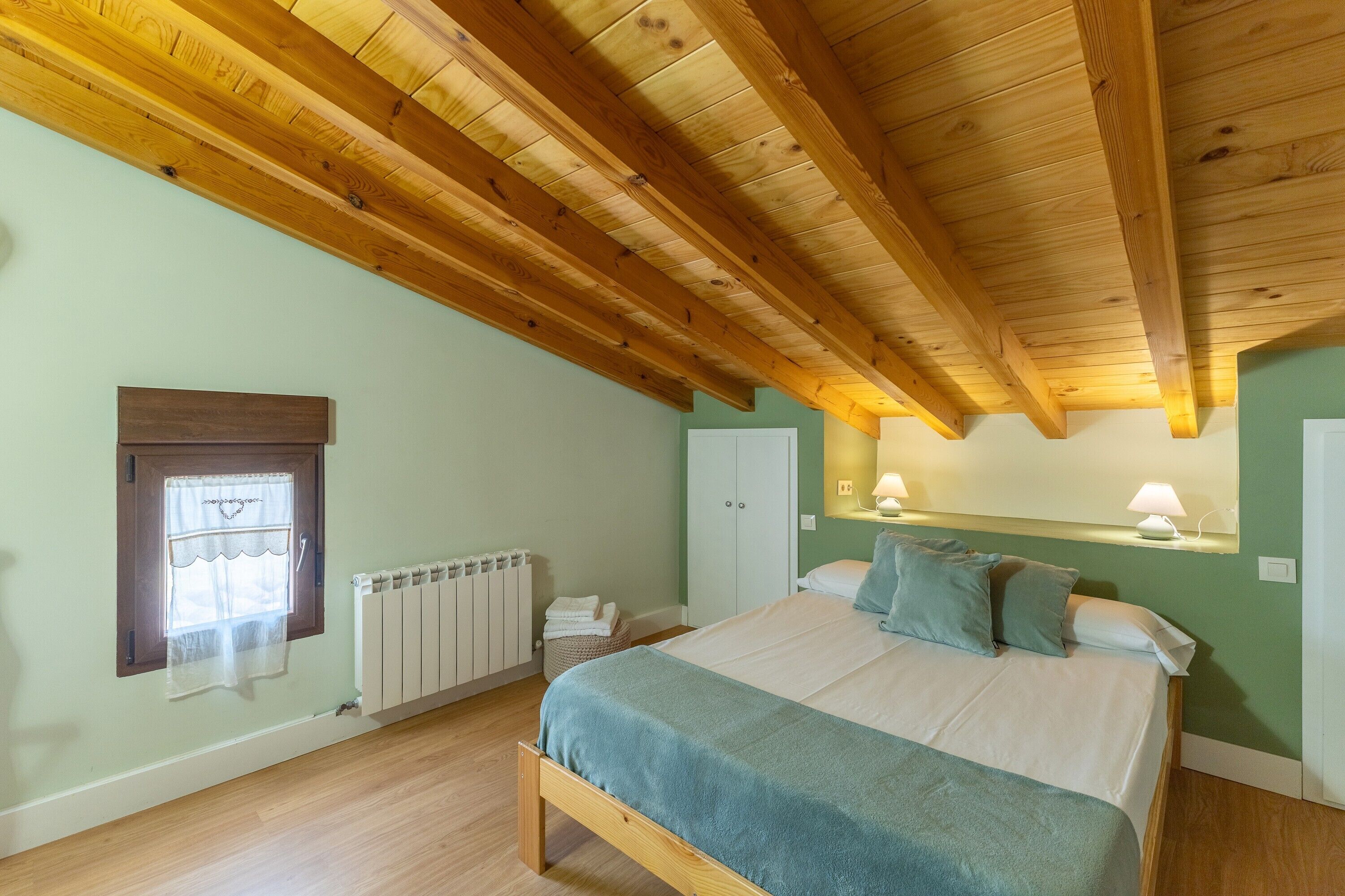 Un dormitorio con techo de madera, una cama con manta azul, un radiador, una ventana con cortina y una lámpara fijada a la pared.