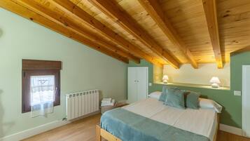 Un dormitorio con techo de madera, una cama con manta azul, un radiador, una ventana con cortina y una lámpara fijada a la pared.
