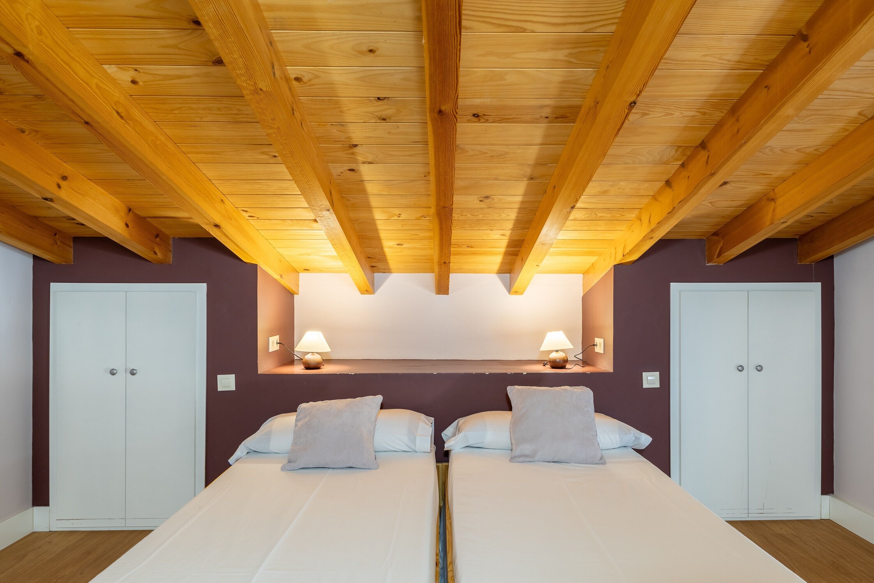 A bedroom with a wooden ceiling, two beds, a headboard, and two bedside lamps.