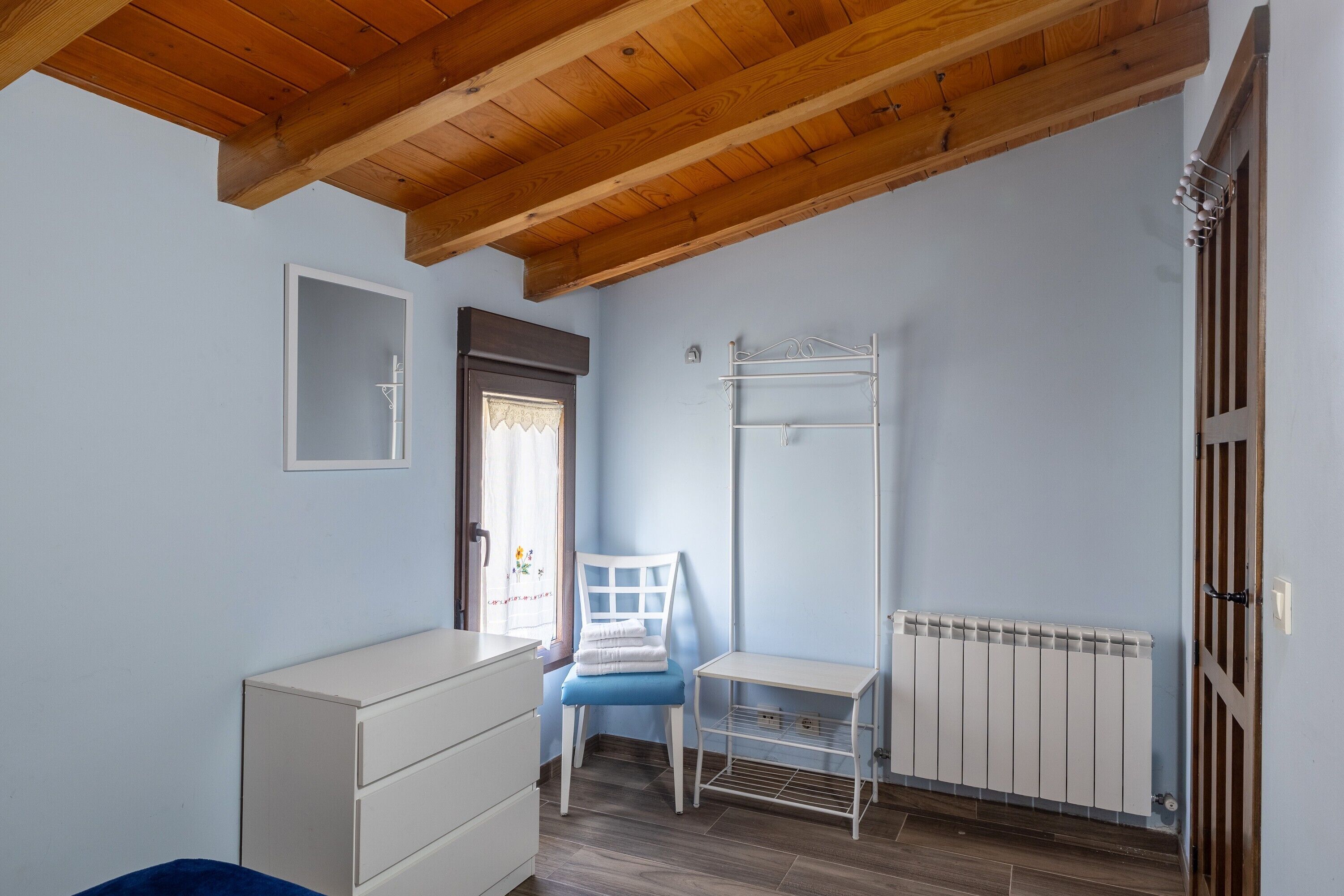 A room with a wooden ceiling, a white radiator, a blue chair, a white dresser, and a window with a curtain.