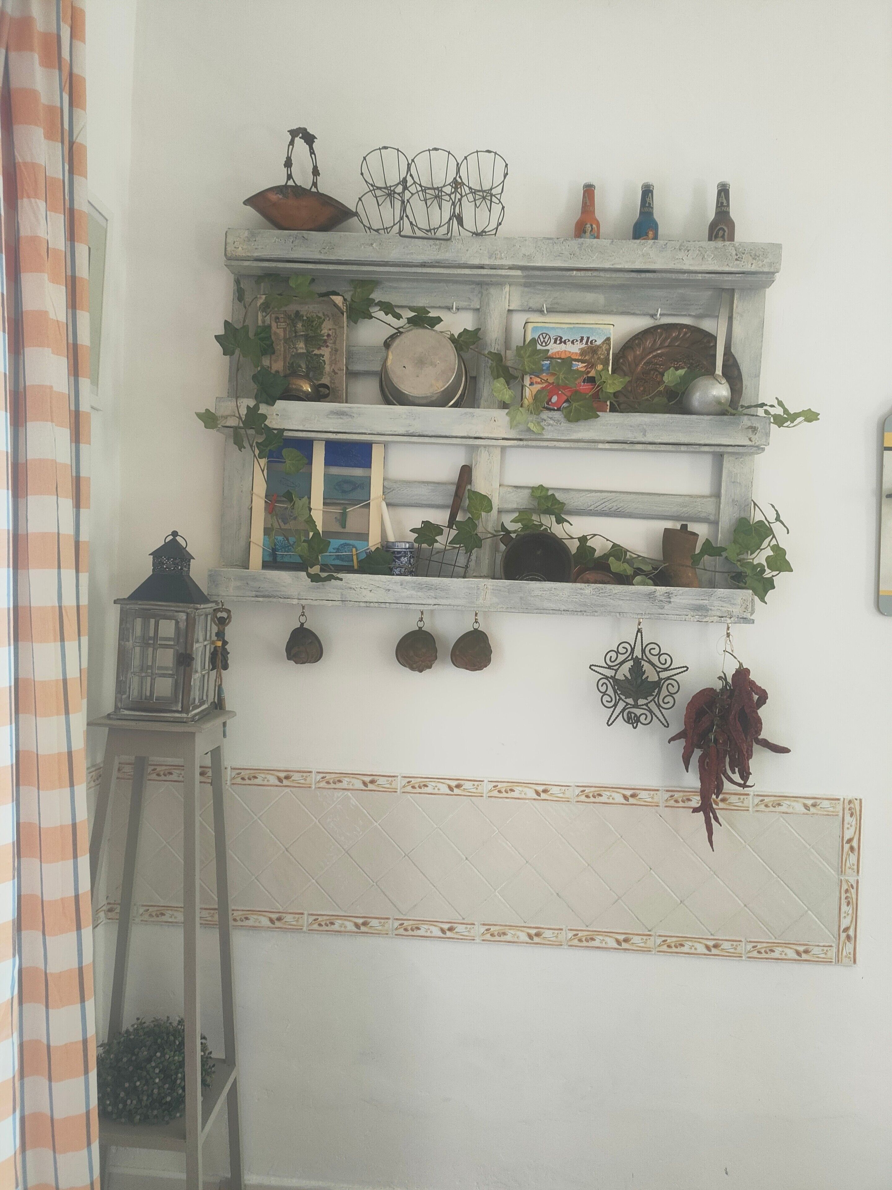Uma cozinha com painel de azulejos brancos, uma escada de madeira, utensílios de cozinha pendurados e uma prateleira com diversos objetos, incluindo uma placa Volkswagen Beetle, uma panela e uma garrafa.