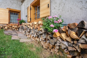 A building with stacked firewood, a stone pathway, and pink flowers in front of wooden windows.