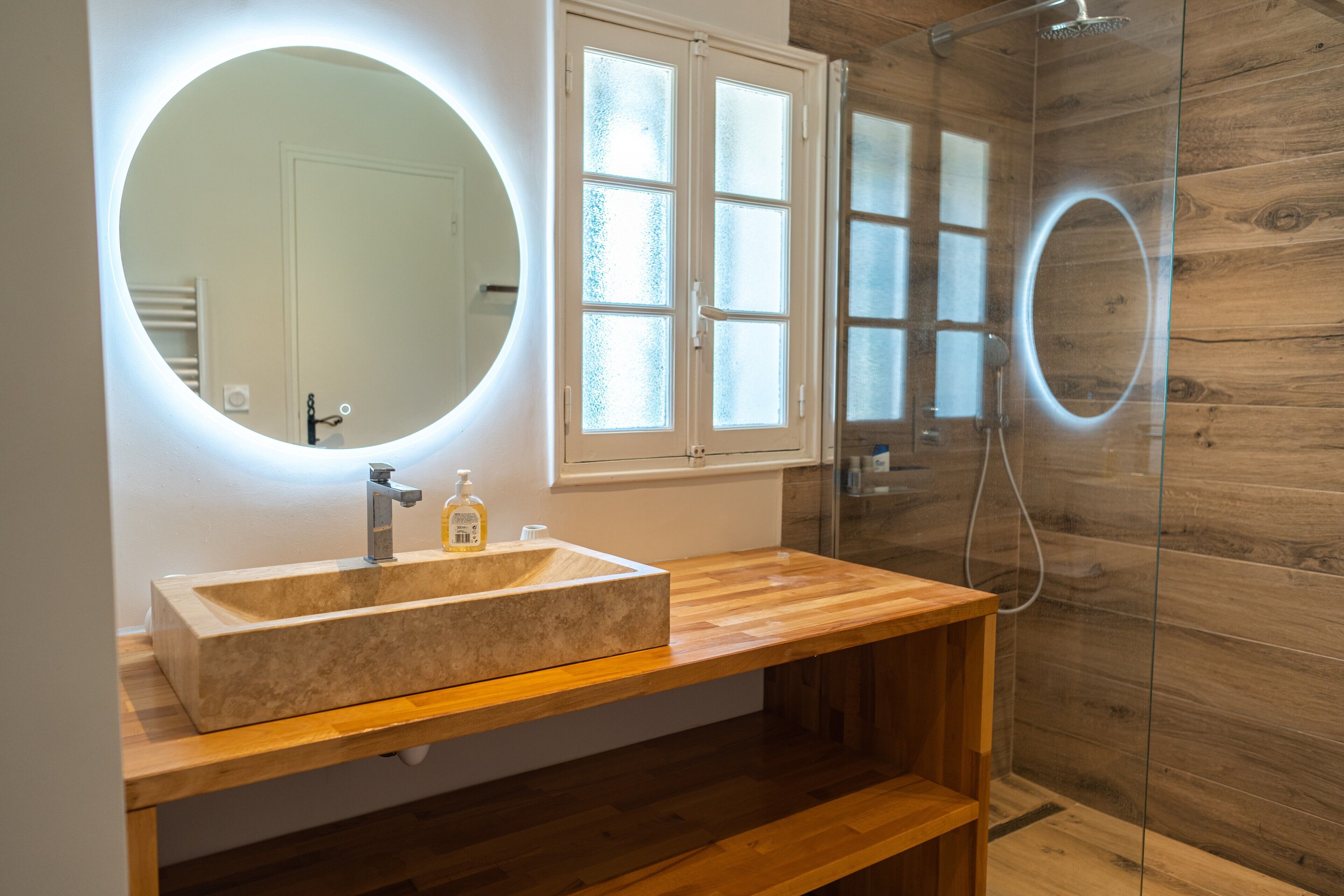 Een badkamer met een houten wastafelmeubel, een grote spiegel met verlichting, een glazen douchecabine en een matglazen deur.