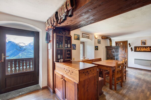 A wooden dining area with a large table and chairs, a wooden cabinet with bottles, and a view of mountains through a glass door.