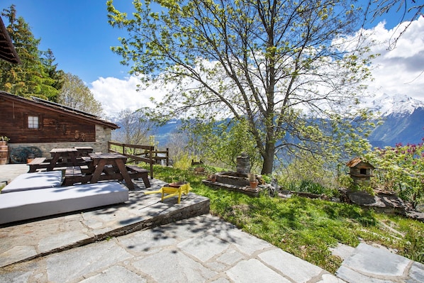 Un chalet rustique en bois, doté d’un plancher en pierre, d’un espace extérieur aménagé et offrant une vue sur des montagnes enneigées.