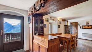 Un coin repas en bois avec une grande table et des chaises, un buffet en bois contenant des bouteilles, et une vue sur les montagnes par une porte vitrée.
