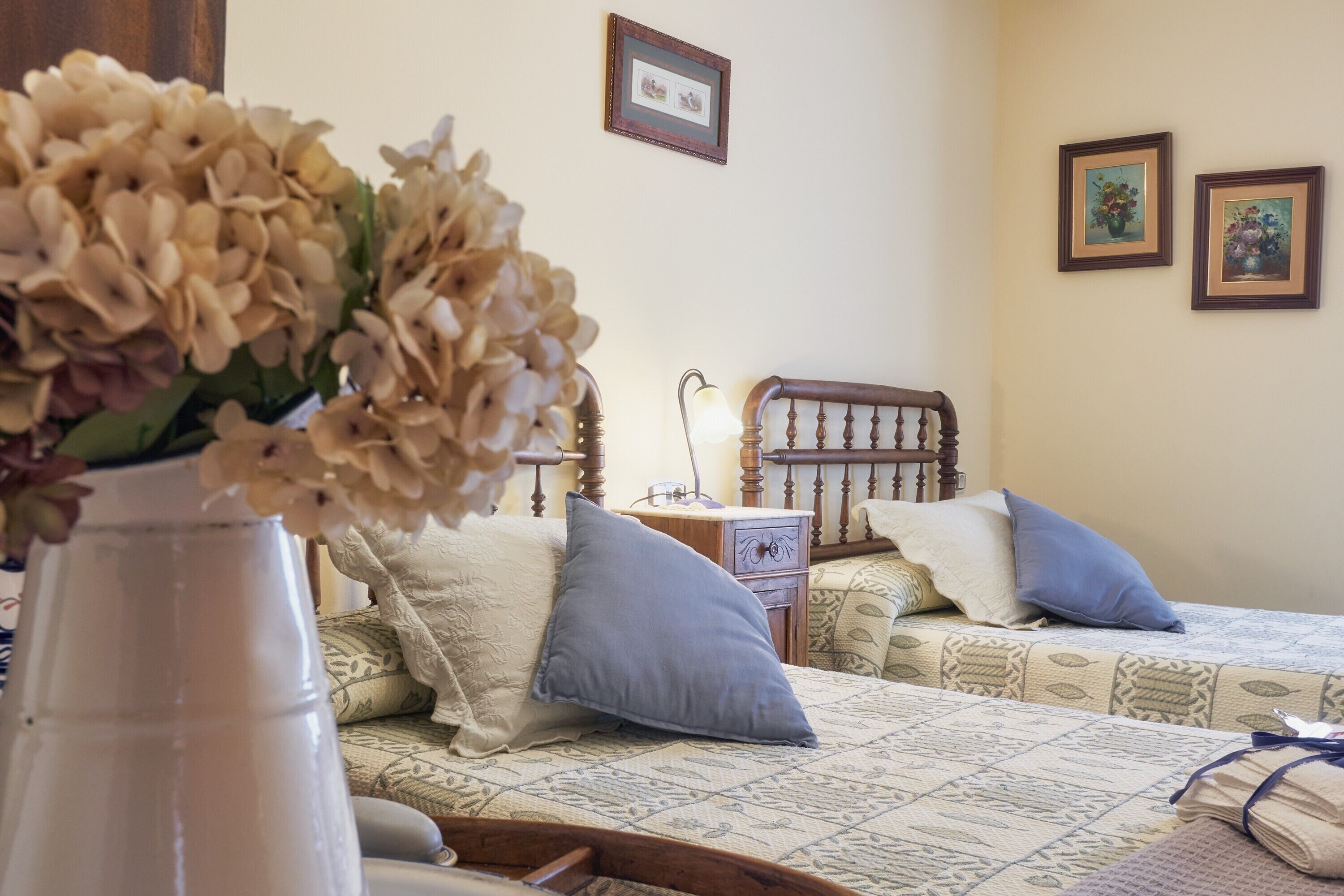 Un dormitorio con dos camas, una mesita de noche, una lámpara y un florero con flores.