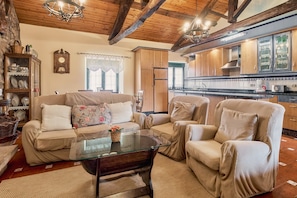 A cozy living room with a sofa, armchairs, a glass coffee table, wooden beams, a chandelier, and a kitchen in the background.