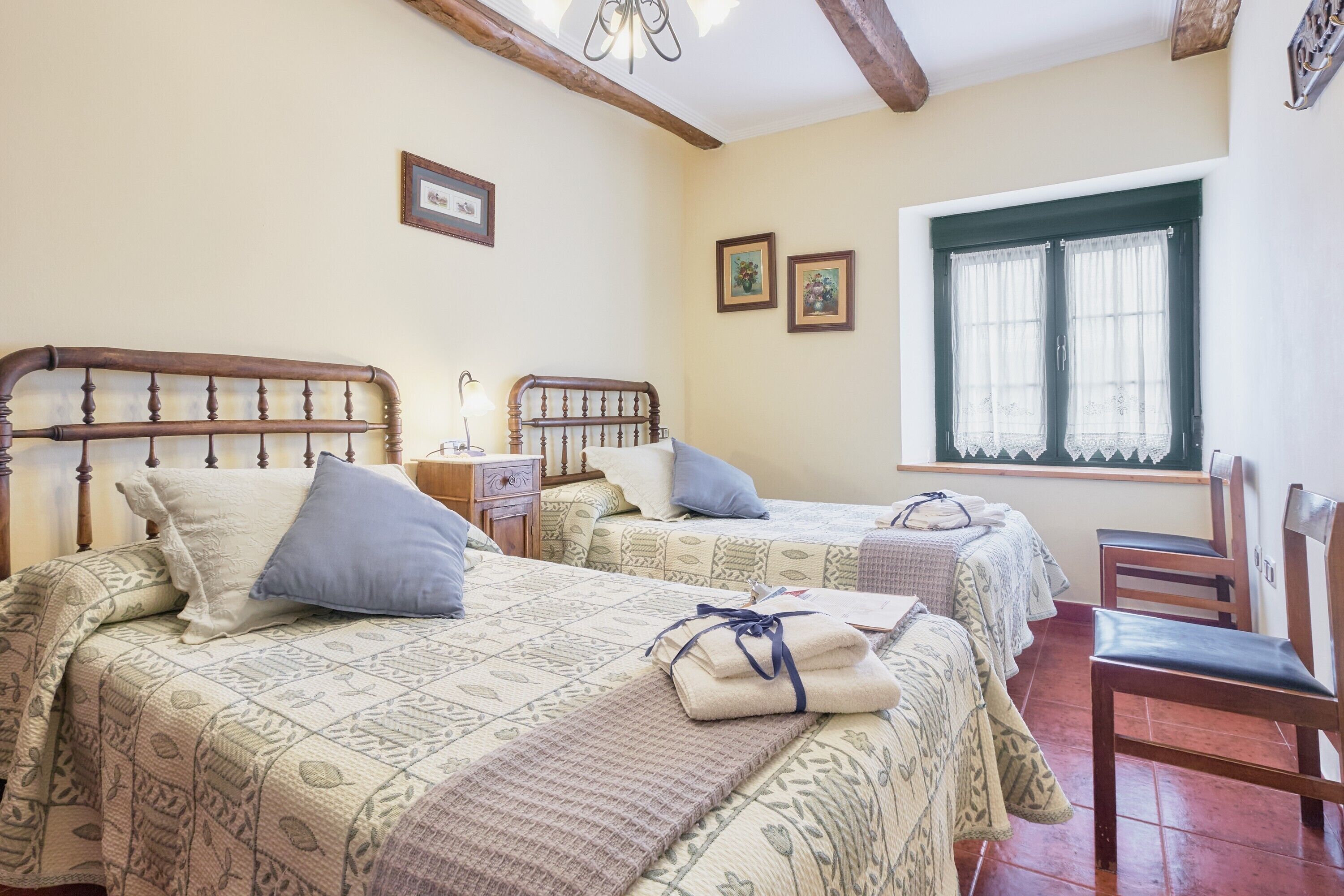 A bedroom with two beds, a window with curtains, a wooden chair, and a nightstand with a lamp.