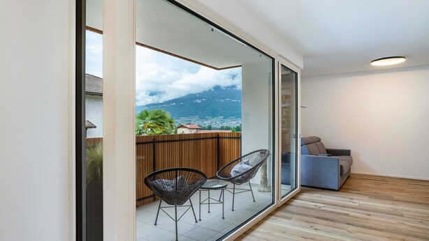 A modern interior with a sliding glass door leading to an outdoor balcony with chairs and a sofa, a wooden floor, and a view of a mountain. - Apartment '2' with Mountain View, Wi-Fi and Air Conditioning (Auer)