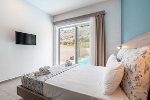 A bedroom with a bed, a TV, a window with curtains, and a view of a pool and hills.