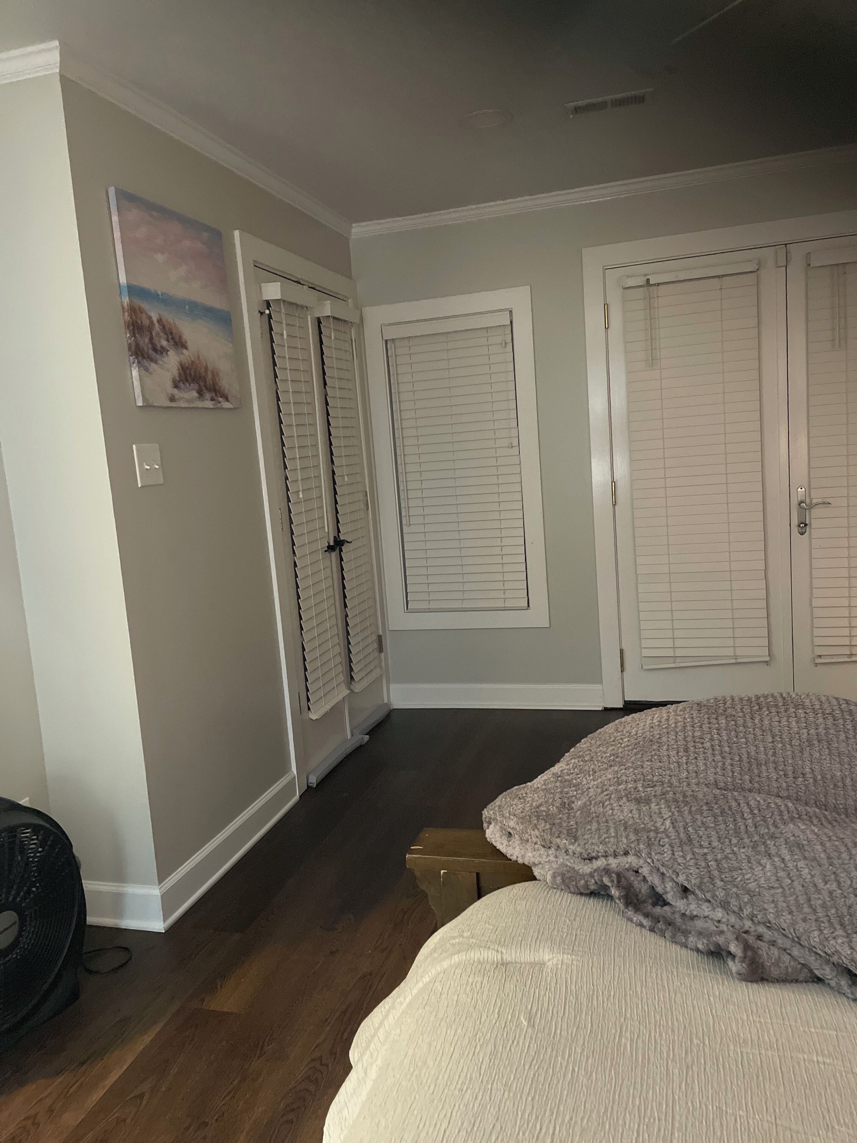 A room with a wooden floor, a large bed, a painting on the wall, and a door with blinds.