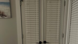 Un juego de puertas dobles blancas con persianas horizontales, piso oscuro y un cuadro en la pared.