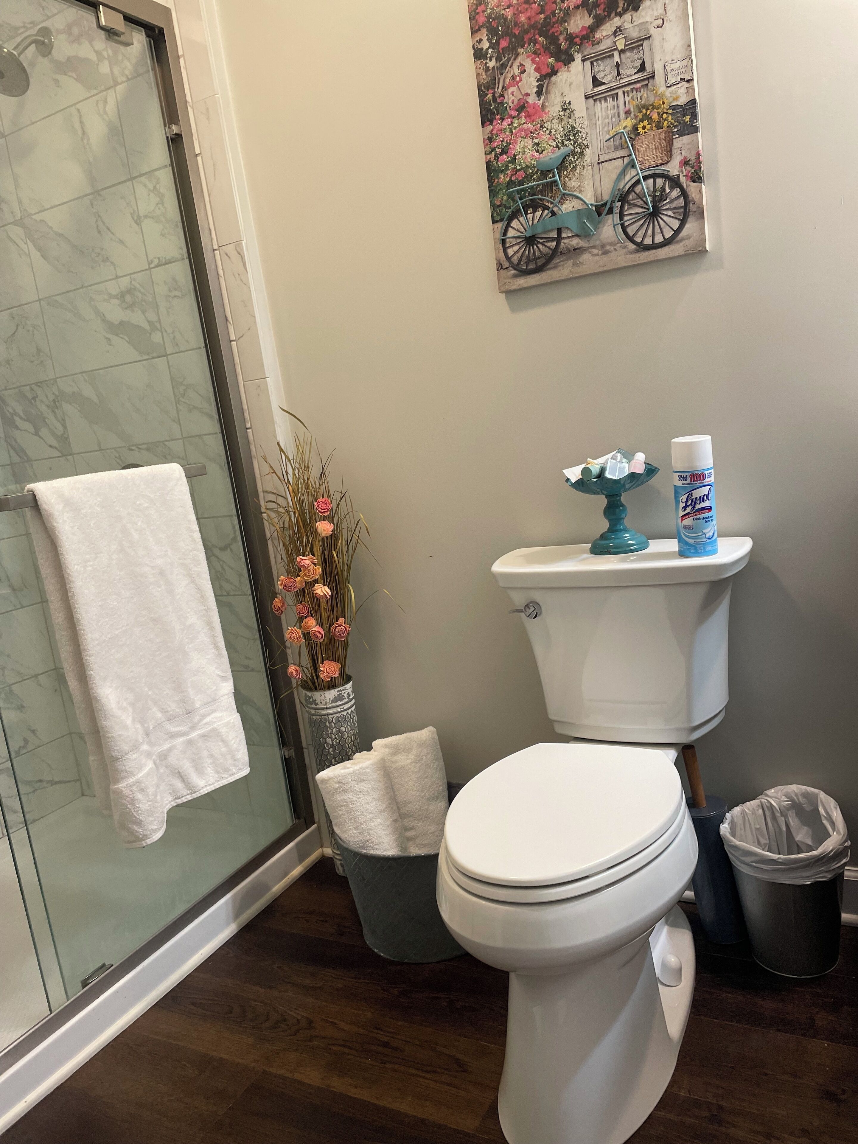 A bathroom with a glass shower enclosure, a toilet, a sink with a decorative blue soap dispenser, a trash can, and a vase with dried flowers.