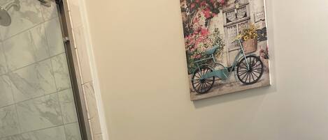 A bathroom with a glass shower enclosure, a toilet, a sink with a decorative blue soap dispenser, a trash can, and a vase with dried flowers.