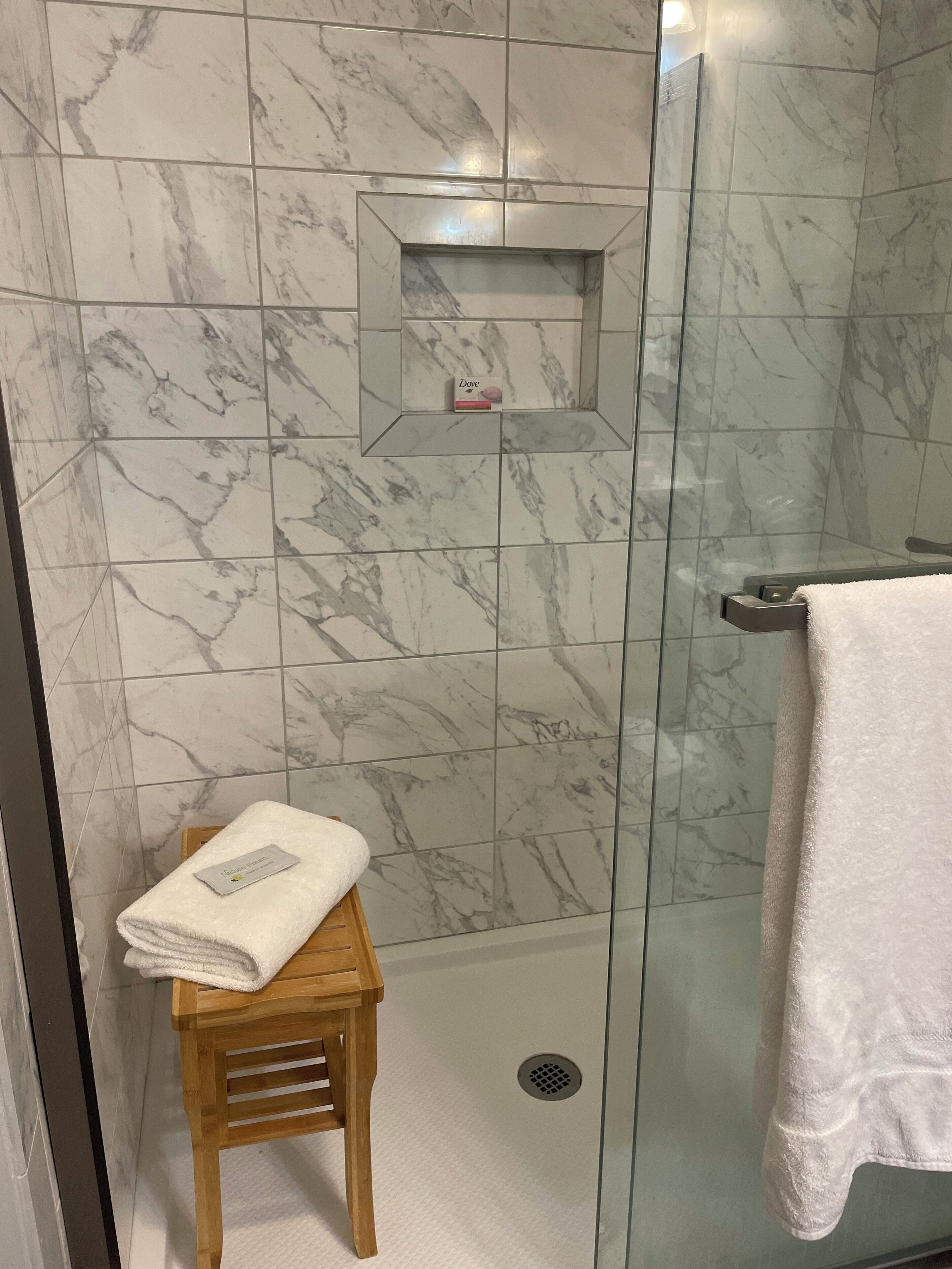 A bathroom with marble tiles, a glass shower enclosure, a wooden stool with a towel, and a towel rack.