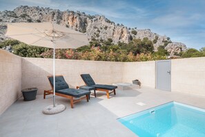 Un espace piscine avec des chaises longues et un parasol, entouré d’un mur de pierre et offrant une vue sur des montagnes rocheuses.