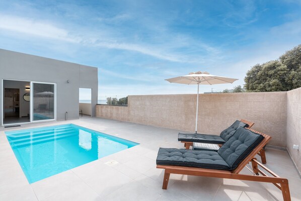 Un espace piscine extérieur moderne, avec une piscine à l’eau d’un bleu limpide, des chaises longues en bois et un grand parasol.