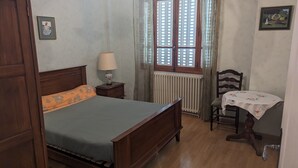 A bedroom with a wooden bed, a nightstand with a lamp, a chair, a small table with a tablecloth, a window with shutters, and a radiator.