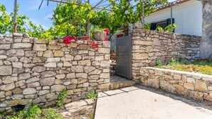 A stone wall with a metal gate, a concrete path, and a building with a tiled roof in the background.