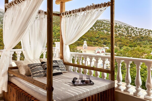 A wooden bed with white curtains, a tray with glasses and cups, and a view of a church and greenery.
