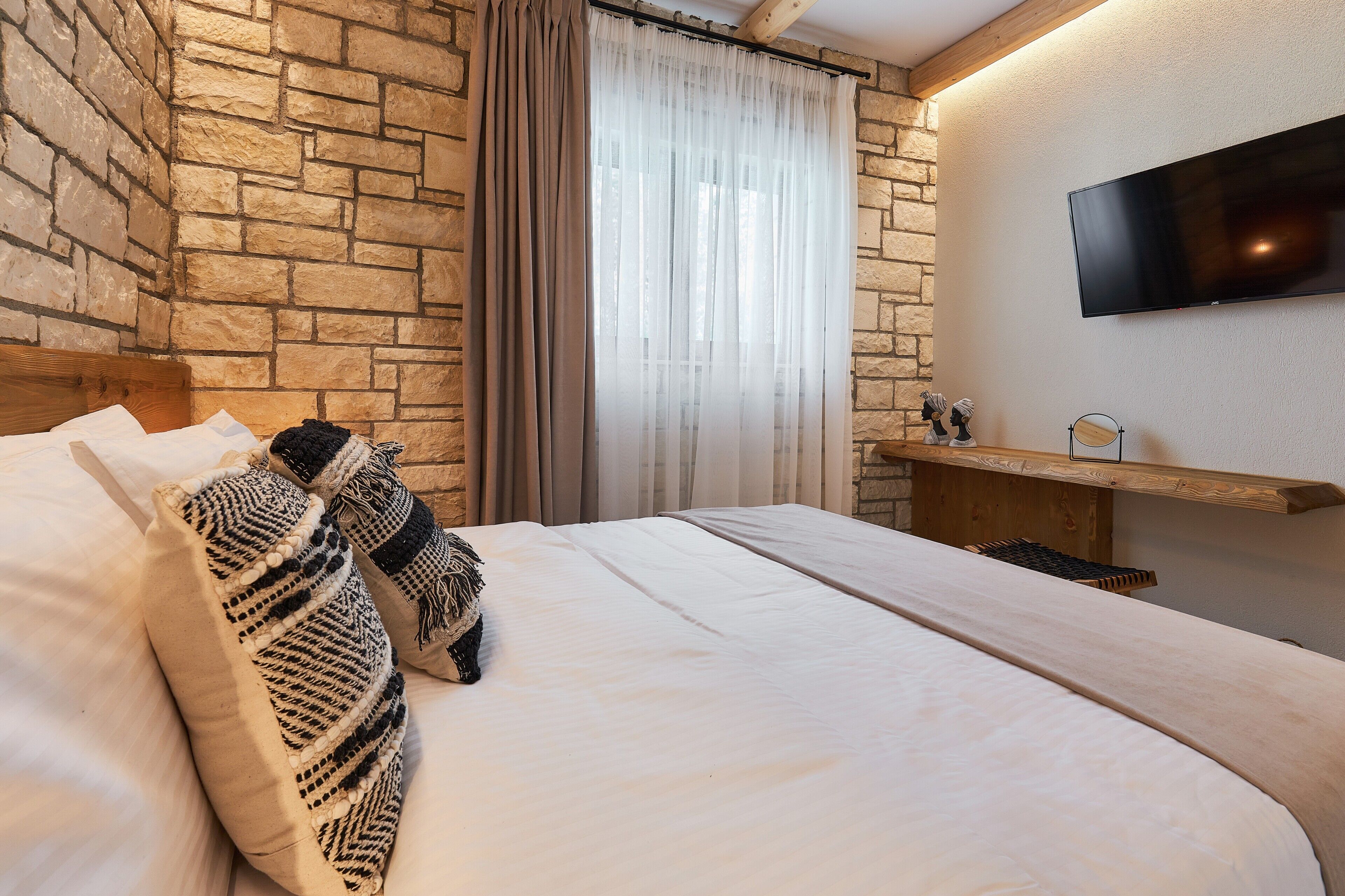 A bedroom with a stone wall, a bed with white bedding, a mounted TV, and a wooden shelf.