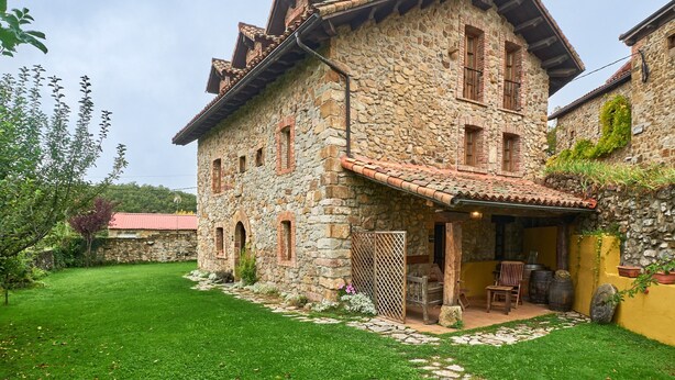 Un edificio de piedra con techo de tejas, un pequeño patio con una silla y mesa, y un camino de piedra que conduce a la entrada.