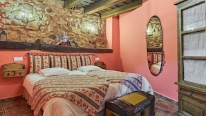 Un dormitorio con pared de piedra, una cama con ropa de cama floral, un cofre de madera y un espejo.