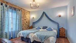 A bedroom with a stone wall, a bed with a blue canopy, two bedside tables, a wooden bench, and a chandelier.