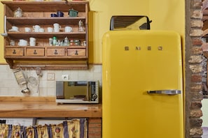 A kitchen with a yellow refrigerator, a microwave, a wooden shelf with dishes, and a brick wall.