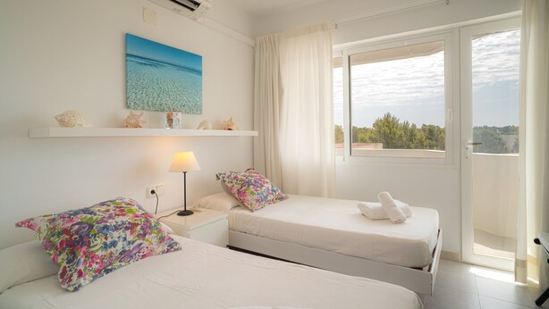 A hotel room with two beds, a window with curtains, a wall-mounted air conditioner, and a painting of a beach scene. - Apartment 'Mirada I' with Open Views, Wi-Fi, and Air Conditioning (Es Pujols)