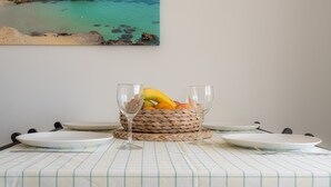 A dining table with a woven basket containing fruit and two wine glasses, set against a beach view painting. - Apartment 'Mirada I' with Open Views, Wi-Fi, and Air Conditioning (Es Pujols)
