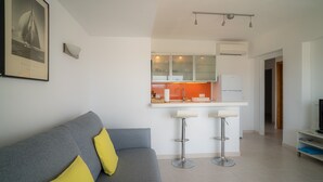 A modern living room with a grey sofa, yellow cushions, a kitchen with white cabinets, and a bar area with stools. - Apartment 'Mirada I' with Open Views, Wi-Fi, and Air Conditioning (Es Pujols)