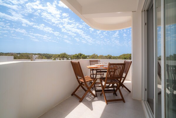 A balcony with a table and chairs, a glass door, and a view of the sky and trees.
