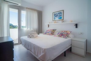 A bedroom with a bed, a nightstand, a dresser, a window with curtains, a wall-mounted shelf with decorative items, and a framed picture.