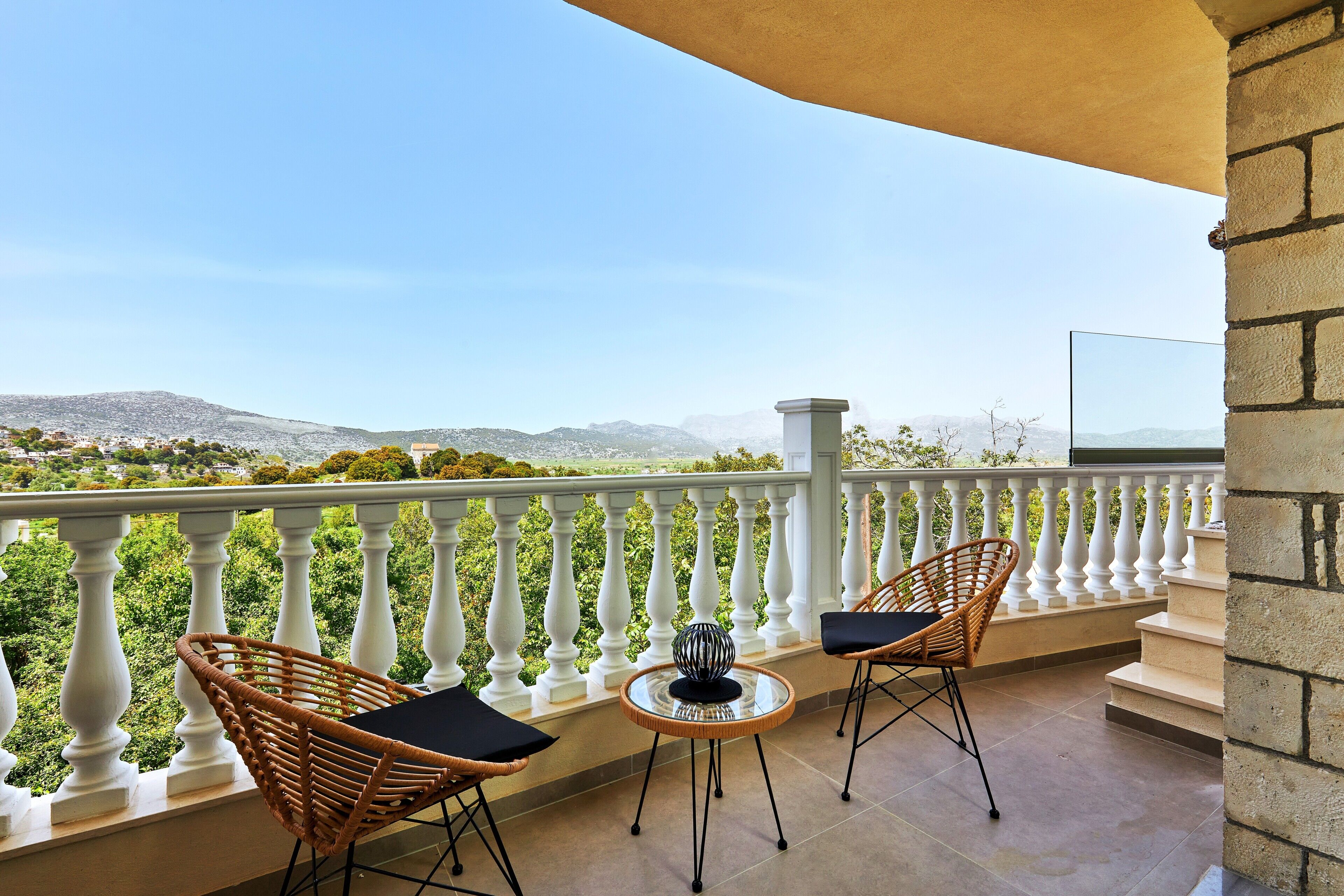 Ein Balkon mit weißen Brüstungen, zwei Korbstühle mit schwarzen Kissen, ein kleiner runder Tisch und eine dekorative Vase.
